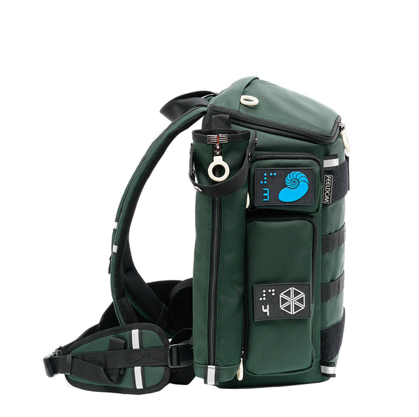 Side view of A forest green boxy backpack with a  green cane pouch attached to the side.  There are braille design velcro patches on the pockets, and a reflective strip across the waist band which is removable by clip.
