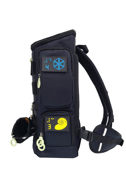 Performance Block Medium is about 6 inches thick, with an upper side pocket and lower access point (double zipper) to the inner compartment. Each access point has a Braille tactile patch. This one shows a blue cube with a number 4 digit and braille, and a yellow nautilus shell with the number 3. The cane pouch is attached to the front molle webbing