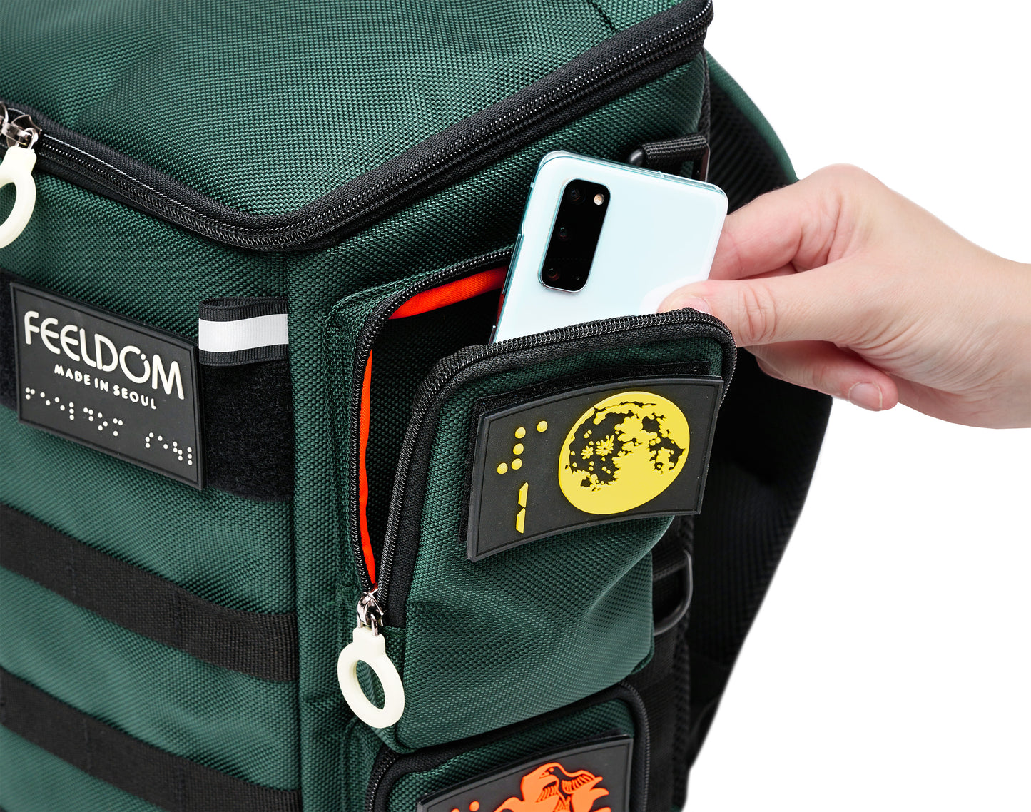 Close up view of the City Block Top left pocket, which is big enough to hold at least 2 smartphones. A person is taking out a smartphone from the pocket, which has a white zipper ring and a braille design patch of the moon and number 1 in braille.