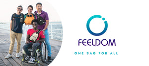 FEELDOM Logo is a blue circle with a dot in the corner. One bag for all. A photo of 4 people standing on a boardwalk wearing Feeldom bags. One guy is visually impaired, and one guy is in a wheelchair. They're smiling
