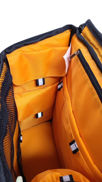 Inside view of the CITY BLOCK backpack, which is a box-shaped structure. The 11 inner pockets are arranged in  various sizes and locations on the top, left, right front and back of the inside of the backpack. They are mede in brignt orange nylon and black mesh, with each pocket having a reflective tab on the opening.
