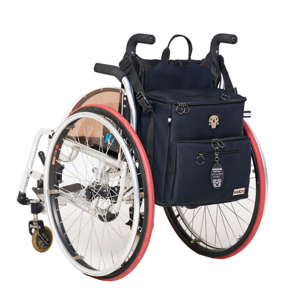 Buddy the Adaptable tote bag is attached to the back of a manual wheelchair. The tote bag has 2 side straps and 2 top straps that clip onto the wheelchair. It has a zip open rectangular lid. It looks like a boxy bag, but cuter. 