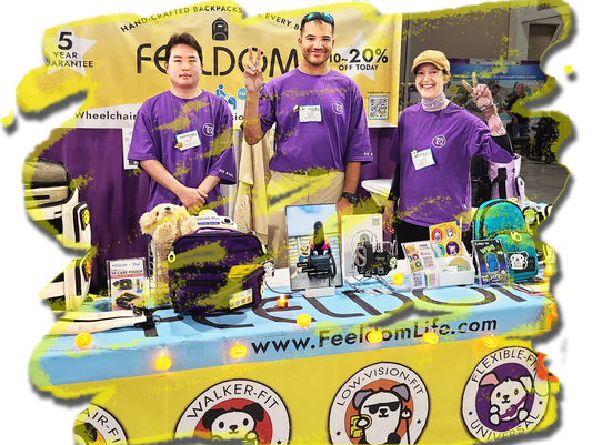 3 smiling people wearing purple FEELDOM logo t-shirts are standing in front of a Yellow FEELDOM banner and there's a yellow table banner with many small items displayed on it.