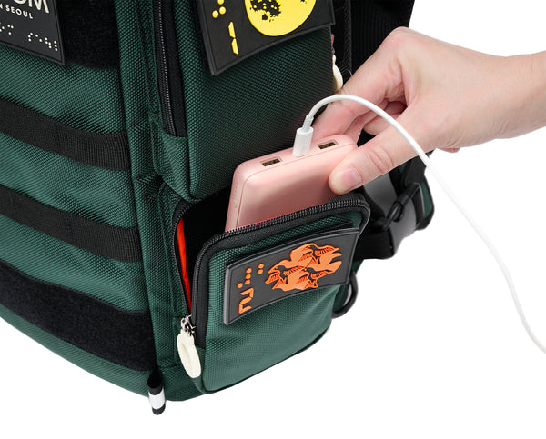 Close up view of the City Block Top left pocket, which is big enough to hold a portable hard drive, battery or wallet. A person is taking out a large battery pack from the pocket, which has a white zipper ring and a braille design patch of orange birds and number 2 in braille.