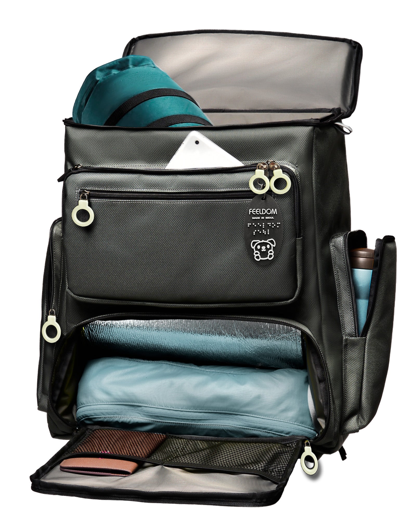 A Feeldom Trek backpack with the box- style flip top lid open, and the lower front panel opened up to the main compartment. In the pack is a small sleeping bag, a camping chair, thermos, passport and cellphone. It has a black and white braille keychain on the front
