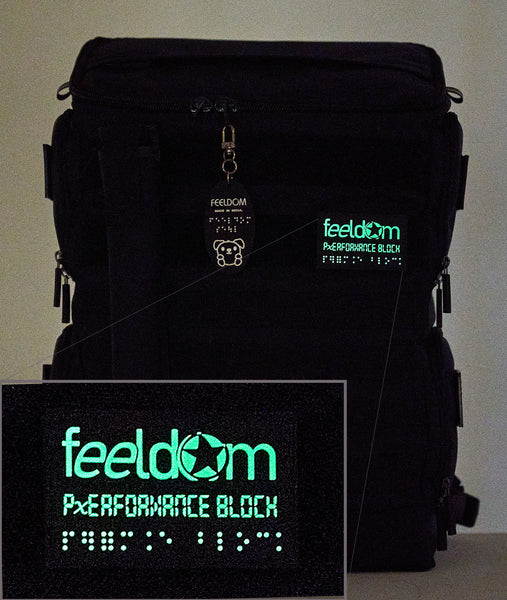 The bag is in the dark, and the Performance Block FEELDOM Logo Braille patch and the P-Block character keychain are glowing in the dark.