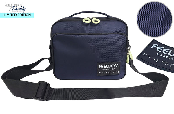 A square shaped travel pouch with a double ring zipper opening on the top and front. There's a Feeldom Logo braille patch on the front lower corner, and a black shoulder strap. Closeup of the Navy blue twill nylon with a diagonal weave. LIMITED EDITION Wheelchair Daddy Collaboration issue