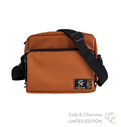 A burnt orange rectangular crossbody bag with a black adaptable strap. There's an embroidered  patch on the front pocket reading CC X Feeldom and an orange puppy in a wheelchair.