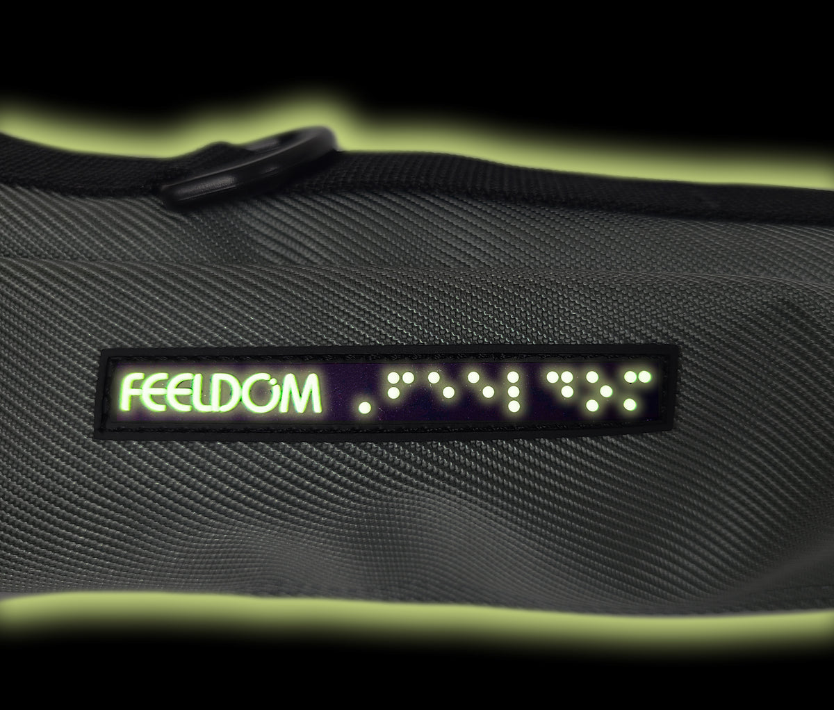 Glow in the dark detail of a small rubber Braille tag sewn onto the side of a pouch. The braille reads FEELDOM next to the logo.