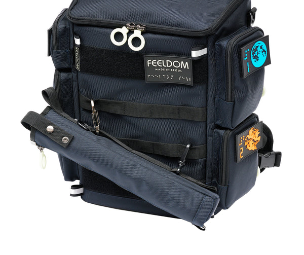 A cane pouch attached to the front of a FEELDOM backpack. The carabiner is attached to the upper webbing strap and the lower clip is attached to the lower strap. The cane pouch is stored diagonally to allow easy access without the can sliding out of the pouch.