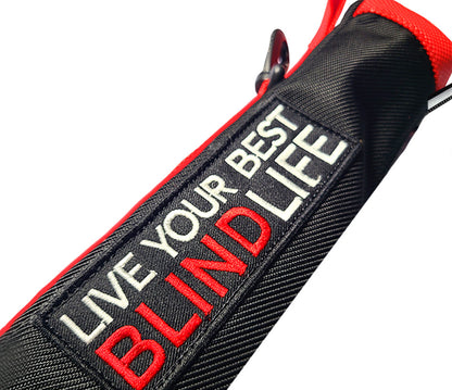 Close-up of a black textured fabric strap or pouch with red trim and a rectangular embroidered patch reading 'LIVE YOUR BEST BLIND LIFE' in white and red text.