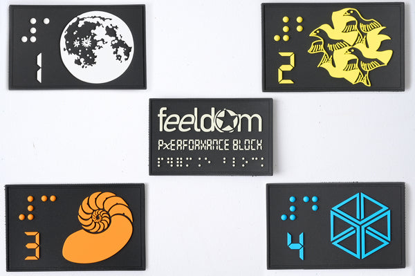 Close up of the Braille tactile patch set of 5. They are black rubber with a raised design in 4 colors. The designs are #1 Moon, #2 Birds, #3 Nautilus, #4 Cube, and the FEELDOM Logo patch . Each one has a braille and number in it. They are velcro on the back.