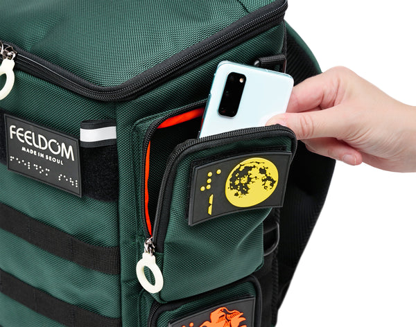 Close up view of the City Block Top left pocket, which is big enough to hold  at least 2 smartphones. A person is taking out a smartphone from the pocket, which has a white zipper ring and a braille design patch of the moon and number 1 in braille.