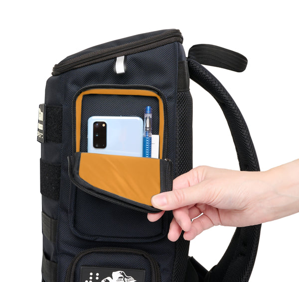 Side view of performance block dark navy medium backpack with an open side pocket showing an orange lining. Inside is a cellphone, a pad of paper and a pen. The rectangle pocket is 5 inches wide, and 7.5 inches tall.