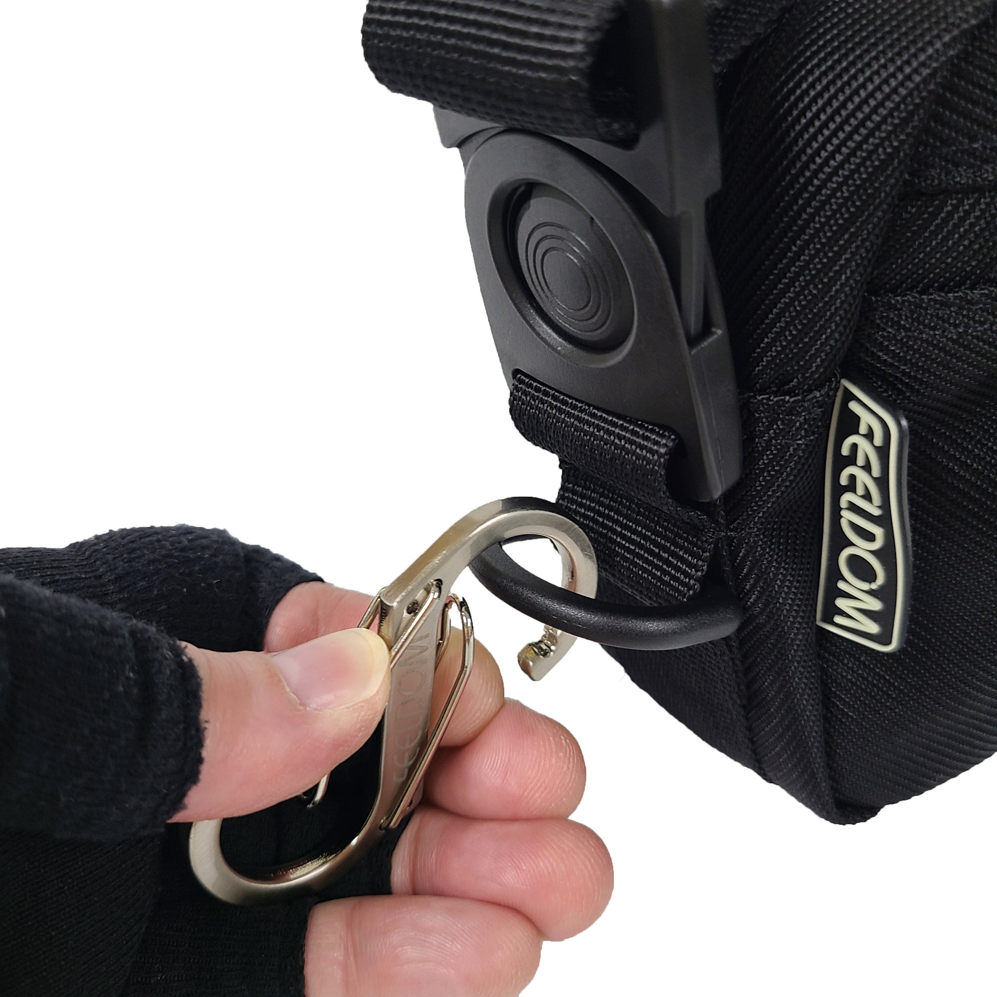 Close up of a metal carabiner being attached to the D ring on the side of a crossbody bag.  The shoulder strap's rotating side buckle clip is also visible.  It has a glow in the dark Feeldom logo tag on the left seam of the bag.