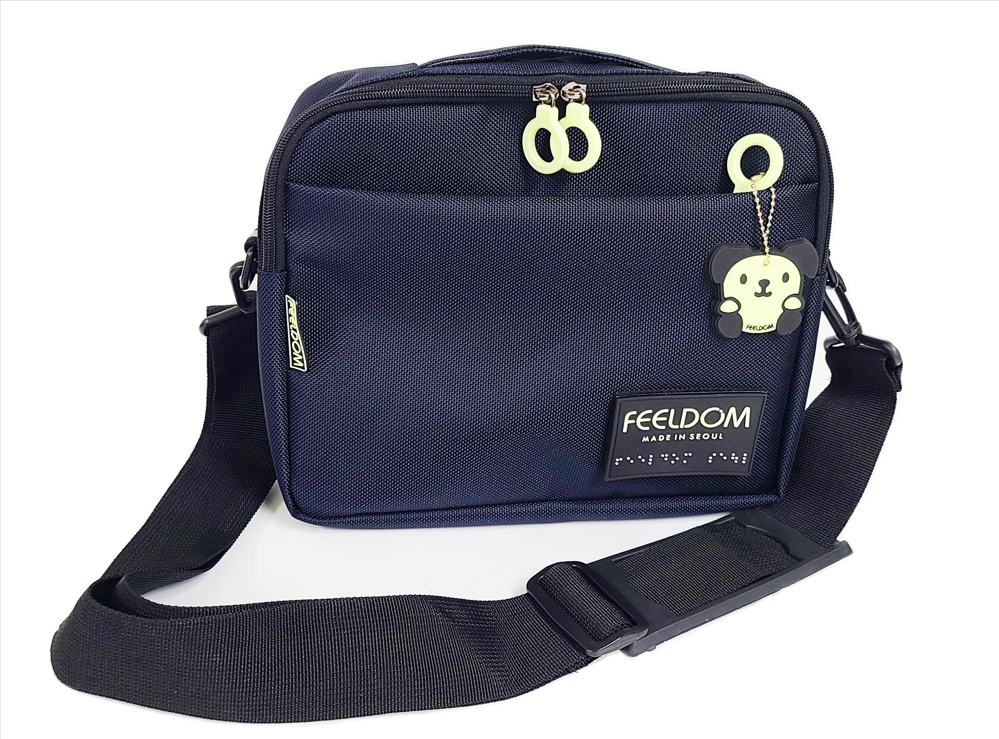 Dark Blue Rectangle pouch with double white zipper closure and front zipper pocket. A glow in the dark dog character rubber keychain and black rubber braille FEELDOM logo patch are on the front. THere is a wide black shoulder strap which is attached to the sides of the pouch. It is 11 inches by 8 inches tall, and 2 to 3 inches thick.