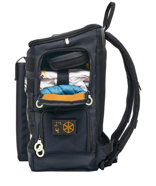 Side view of Perfromance Block HD-23 Large backpack with the upper compartment open from the side. In there are a headphones case, a folded martial arts uniform and a down vest. There are two openings on the side, each with a braille design patch and double white zipper rings. The lining is bright orange.