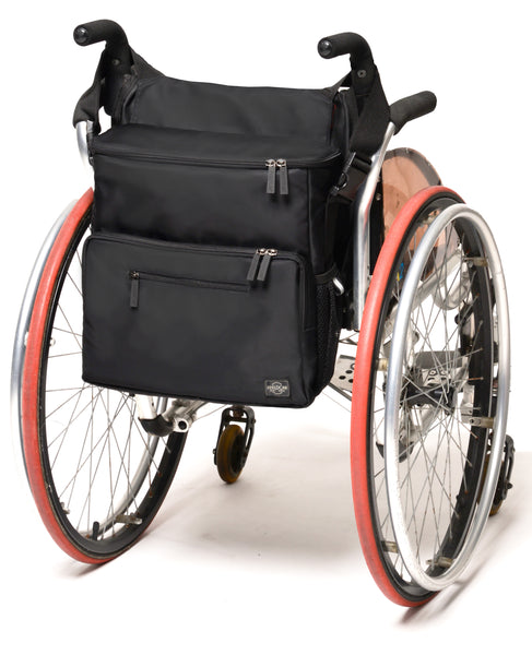 Chic Adaptable Totebag is on the back of a manual wheelchairs. The straps are looped around the handles and the bag is square and hangs down with a slim profile. It is about 12 inches by 12 inches square, with a 4 inch total width.
