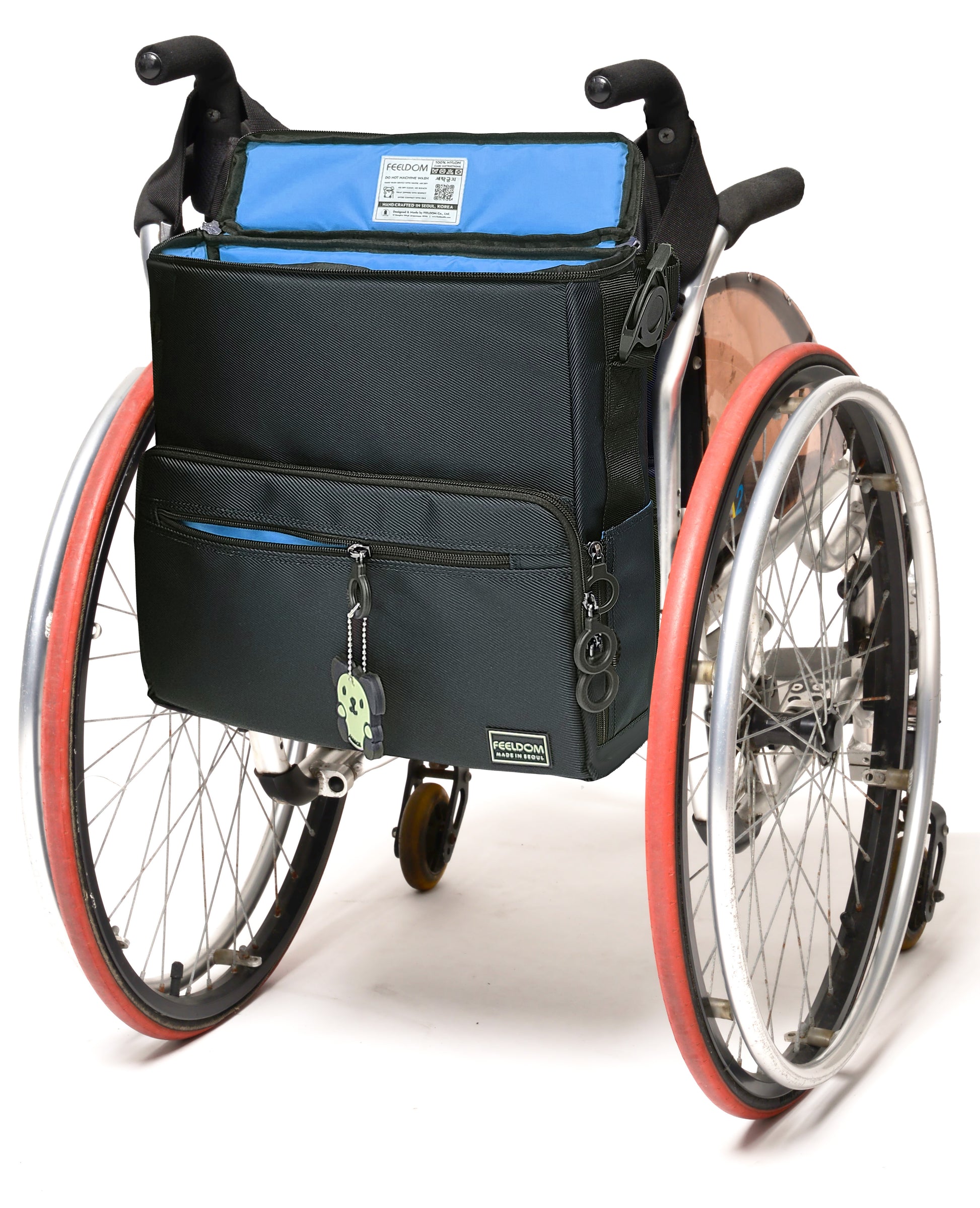 A square -shaped bag attached to the back of a manual wheelchair. It is navy blue with a sky blue lining. It has zipper side pockets that can hold a container. The clips that attach the straps are rotating push botton buckles, like a seatbelt