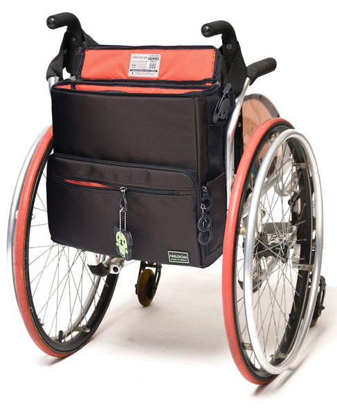 A square -shaped bag attached to the back of a manual wheelchair. It is dark chocolate brown with a watermelon red lining. It has zipper side pockets that can hold a container. The clips that attach the straps are rotating push botton buckles, like a seatbelt