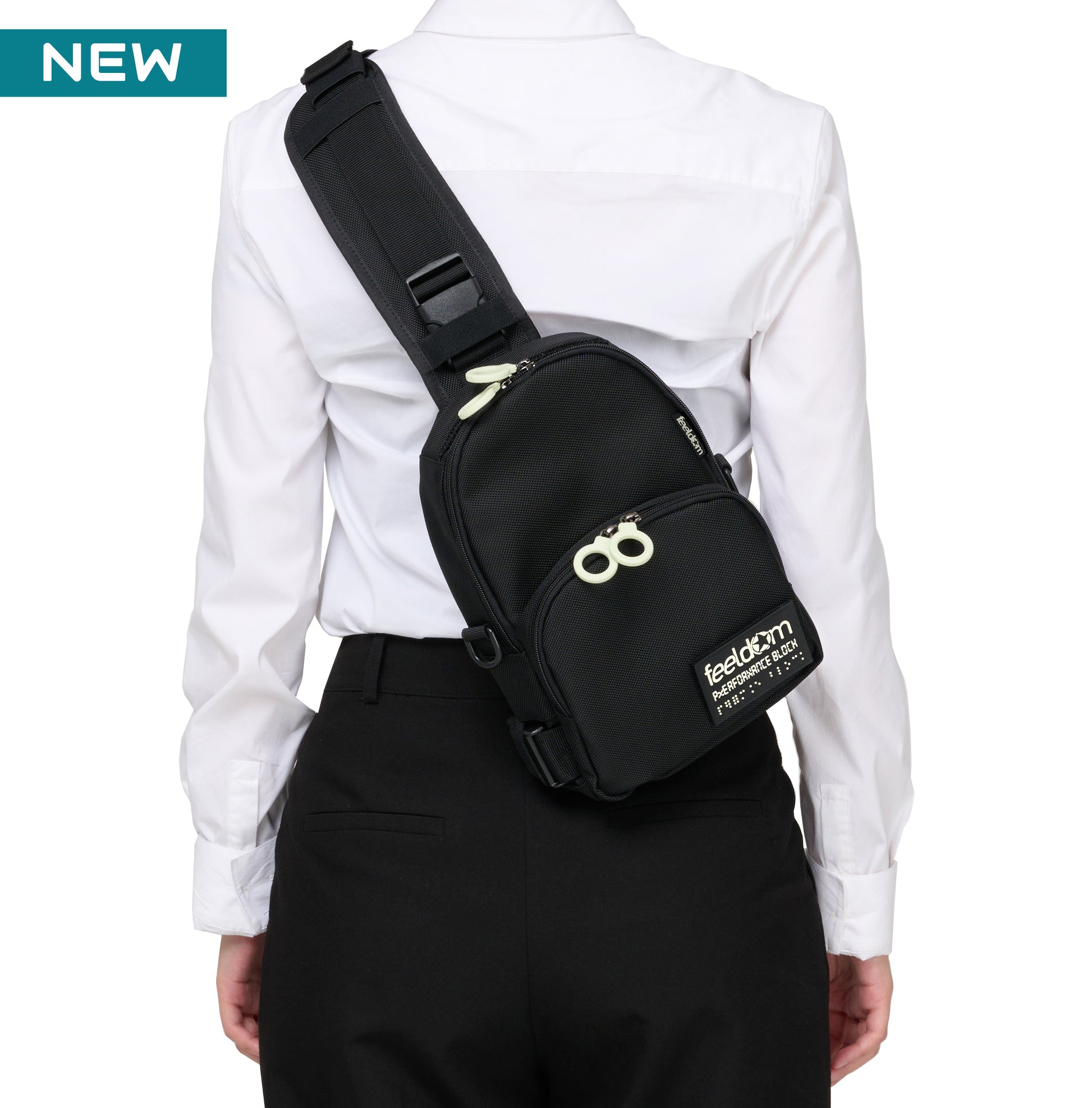 A person wearing a black cross body bag along their back. It has white zipper rings and a black and white Feeldom Braille label.