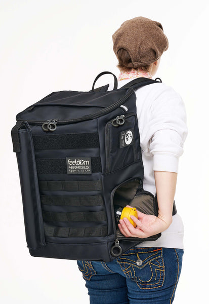 A person wearing the Performance Block Large on their back. They are reaching back with their right hand to take out a thermos from the lower compartment. It looks easy to get, because of the ring zippers and large side-access points. Performance Block L really looks like a big block, and it has black velcro strips and MOLLE webbing on the front, as well as a cane pouch and glow-in-th-dark Feeldom Braille patch.
