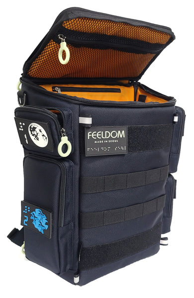 A Dark Navy, box-shaped backpack with black velcro and black straps, colorful braille patches on the side and an orange lining. The flip-top lid is open to reveal an inner pocket with a white zipper ring. Label reads FEELDOM Seoul in glow-in-the-dark Braille.
