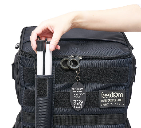 Performance Block has a nylon can pouch attached to the front with velcro. A hand reaches out to take out the white cane out of the pouch. The premium black plastic ring zippers say "Feeldom" on them, and the glow in the dark patch reads "Feeldom Performance Block" in Braille