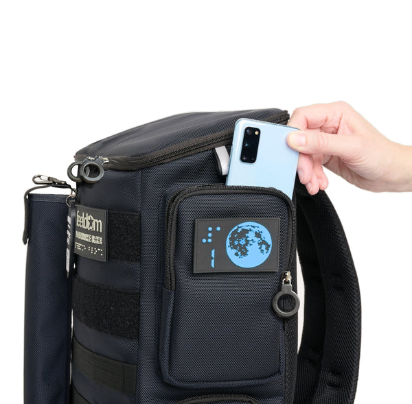 a hand is taking out a cell phone from the upper side pocket of perfromance block medium. The pocket has a blue Moon design. The bag is navy blue and has black ring zippers.