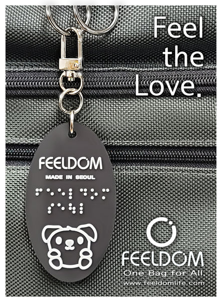 Goldie mascot black rubber keychain with white design is hanging from the rings of a wheelchair bag. Caption reads "Feel the Love"