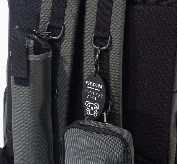 Back view of a gray backpack and black air mesh back padding. The padded straps have 3 D rings on each one. Attached to the D rings are a gray cane pouch, a small phone case pouch, and a braille keychain with a dog character on it.