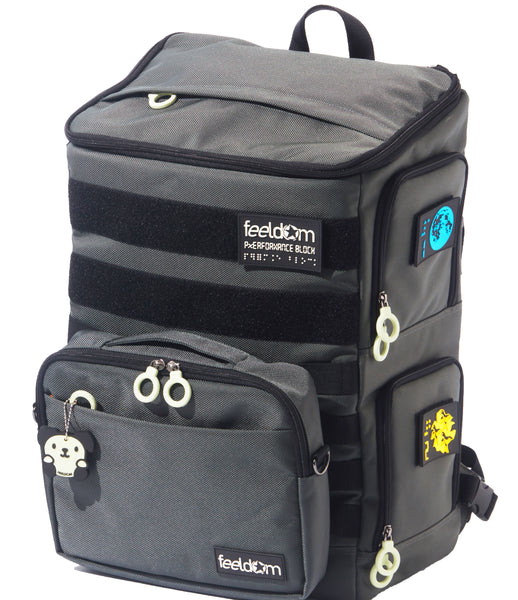A large backpack with side openings. Gray ballistic nylon with white zipper rings and bright braille patches on the side. There is a zipper on the top with a rain cover. There is an additional pouch attached to the front. This TEKNO Pouch is sold separately. Glow in the dark labels read FEELDOM.