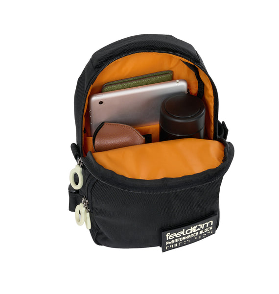 Open view of a black crossbody bag with a bright orange inner lining.  Inside the larger main compartment is a small ipad, a passport, a thermos and sunglasses. The opeing folds down to the center of the bag allowing quick access. There is a padded divider in there to keep the ipad safe from scratches