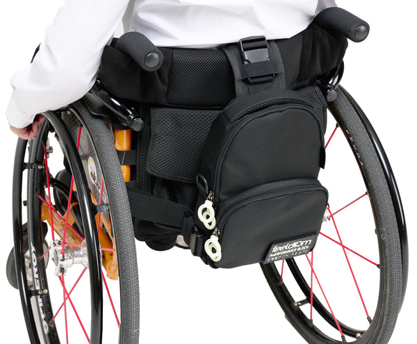 A black crossbody bag is attached to the back of a manual wheelchair with the large main strap going up and over the back of the seat back. The long horizontal strap runs completely around the seat and is clipped to both sides of the lower part of the bag.