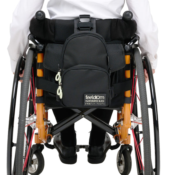 A black crossbody bag is attached to the back of a manual wheelchair with the large main strap going up and over the back of the seat back. The long horizontal strap runs completely around the seat and is clipped to both sides of the lower part of the bag.