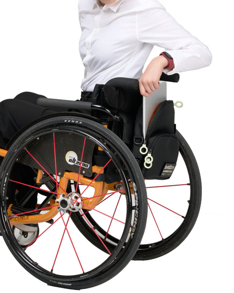 A black crossbody bag attached to the back of a manual wheelchair. The person is taking an i pad out of the bag.