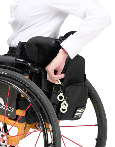 A black crossbody bag attached to the back of a manual wheelchair. The person is reaching back to take hold of the side ring zippers.