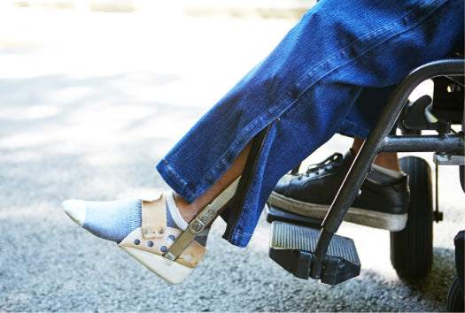A view of a person with a leg brace seated in a wheelchair. The leg brace is visible because the Real Jeans BraceX are zipped open from the bottom to the middle calf.
