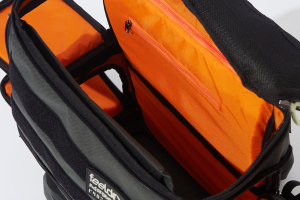 Inside view of a Large backpack with bright orange lining and a divider for the upper and lower compartments. Side openings are on the right and left.  The top lid opens for a 13 inch Laptop computer and under the lid is a mesh zipper pocket.