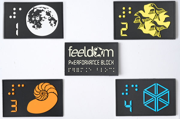 A display of 5 black rubber tactical patches. They are rectangular, 4 x 7 cm. Each one has a different color raised design with braille numbers and digital numbers. White moon is number 1. Yellow birds are number 2. Orange Nuatulus is number 3. Blue 3-D cube design is number 4. The middle patch is our Feeldom logo with the digital text reading "Performance Block." Also written in Braille. All patches are removoable velcro.