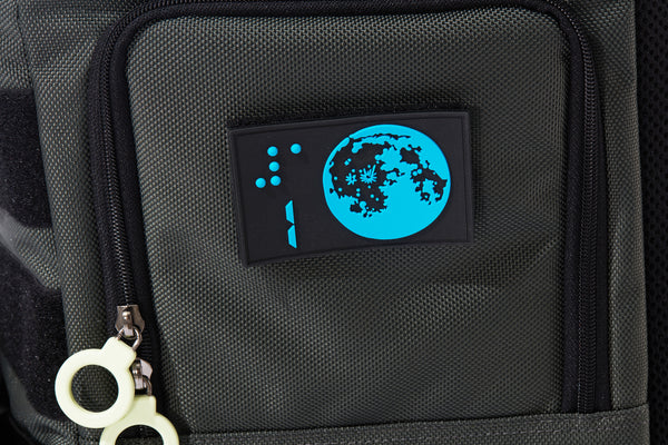 A close up of the side of the large gray backpack.  There is a rectangle rubber patch with the design of the moon and braille number one. It is blue on a black background.