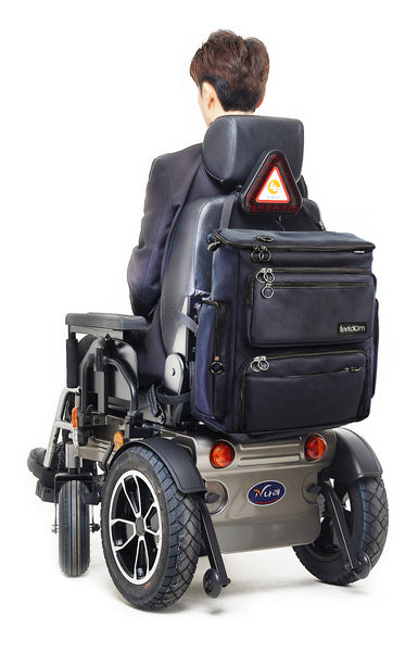 Back view of an electric wheelchair with the Feeldom Max Large Wheelchair bag on the back. It is boxy, dark navy ballistic nylon with metal ring zipper pulls.