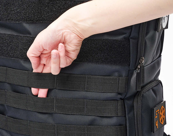 A close up of the front of the tactical backpack in dark navy shows a hand reaching toward the middle.  Two fingers are being slid behind the tactical straps to indicate that they are functional straps perfect for clipping and attaching gear and accessories. 4 of these strips run horizontally across the lower part of the front. The upper part of the bag has 2 wide strips of black velcro for the attachment of patches and gear as well.
