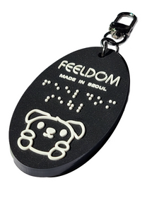 Feeldom's mascot, Goldie the service pup, is the character on our Black rubber oval-shaped keyfob. The white lettering "Feeldom, made in Seoul" are also in Braille (white). It has a 2-inch dark nickel chain and U-shaped clip for versatile use. 