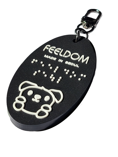 Feeldom's mascot, Goldie the service pup, is the character on our Black rubber oval-shaped keyfob. The white lettering "Feeldom, made in Seoul" are also in Braille (white). It has a 2-inch dark nickel chain and U-shaped clip for versatile use. 