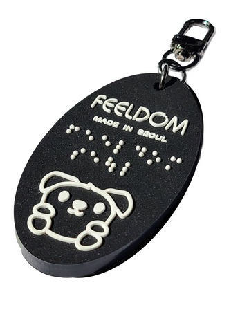 Feeldom's mascot, Goldie the service pup, is the character on our Black rubber oval-shaped keyfob. The white lettering "Feeldom, made in Seoul" are also in Braille (white). It has a 2-inch dark nickel chain and U-shaped clip for versatile use. 