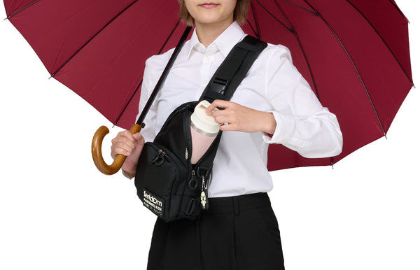 A person carrying the JAYU black crossbody bag on their front while holding an umbrella with one hand.  With their other hand, they are removing a small thermos from the bag.