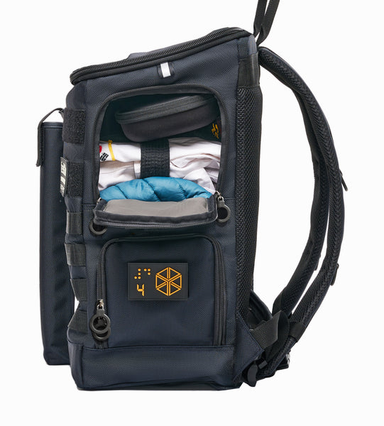 Side view of Navy Blue with open upper level side compartment. Inside is a Taekwondo uniform, down vest, and headphone case. Lower level is zipped closed with double ring zippers. Padded backpack straps and waist belt are black with D rings to attach accessories. Reflective tabs on the sides. 5 inch black handle at the top.